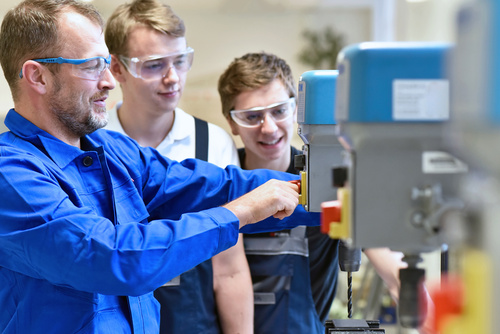 Lehrherr und Lehrlinge in Arbeitskleidung an einer Standbohrmaschine