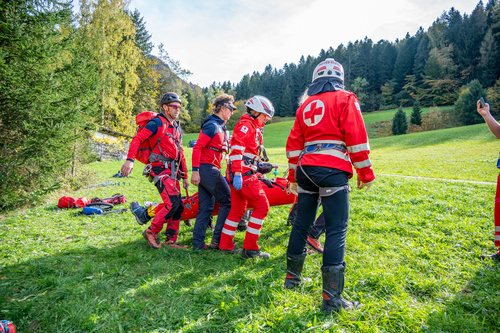 Bergrettung, Feuerwehrleute und Personen des Rettungsdienstes auf einer Wiese beim gemeinsamen Tragen einer Person in einer Trage