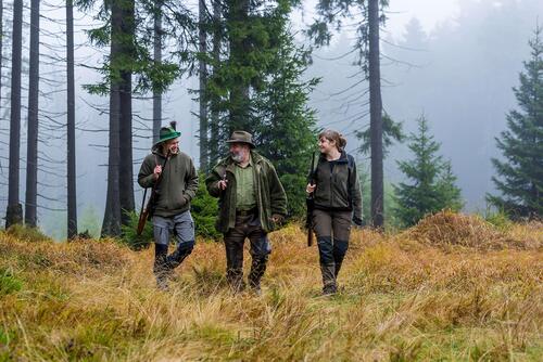 Zwei Männer und eine Frau in Jagdbekleidung in einem Wald.