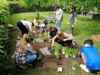Neun Jugendliche pflanzen in einem Garten Kräuter in die Erde, daneben stehend eine Trainerin mit zwei weiteren Kräutertöpfen in Händen