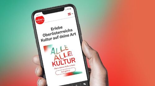 Hand hält Smartphone, auf dem Bildschirm ist die geöffnete Website des OÖ Tourismus mit der neuen oö. kulturtouristischen Kampagne mit dem Titel „Alle Alle! Kultur“ zu sehen.
