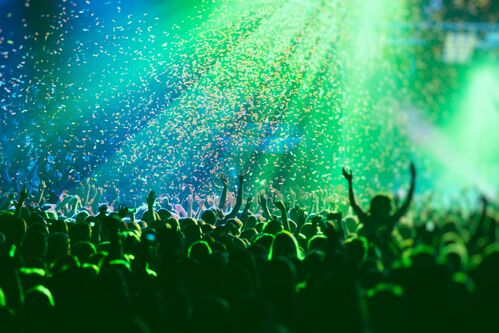 Ein überfüllter Konzertsaal mit grünen Lichtern auf der Bühne, Rockshow-Performance, mit Menschensilhouetten, bunte Konfetti-Explosion, die auf der Tanzfläche abgefeuert wird.