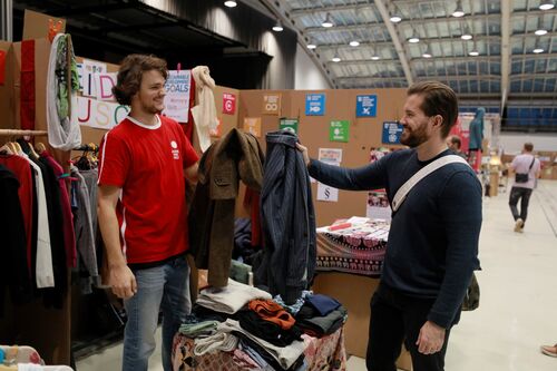 Zwei junge Männer stehen sich in einer Halle gegenüber und halten beide ein Kleidungsstück in die Höhe, neben ihnen Verkaufsstand mit Kleidung. 