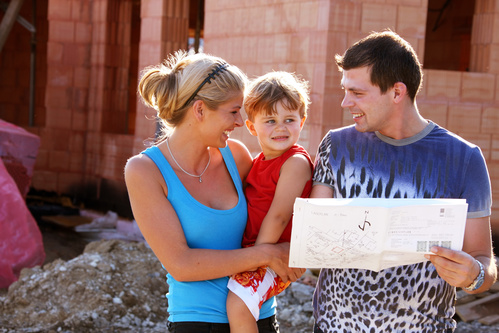 Frau mit Kind auf dem Arm und Mann mit Bauplan in Händen vor dem Rohbau eines Hauses