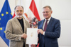 Prof. Mag. Dr. Alfred Weidinger und Landeshauptmann Mag. Thomas Stelzer stehen nebeneinander und halten gemeinsam ein Dokument, im Hintergrund EU- und Oberösterreich-Fahne