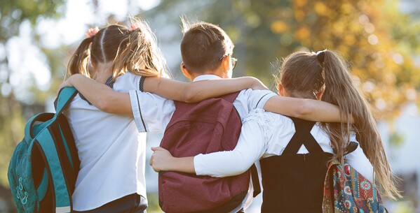 Drei Kinder mit Schulrucksäcken haben die Arme um die Schultern des jeweils anderen gelegt und gehen gemeinsam in eine Richtung