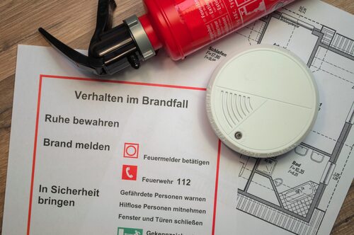 Auf einem Tisch liegen ein kleiner Feuerlöscher, ein Rauchmelder, ein Wohnungsplan und eine Infoblatt Verhalten im Brandfall